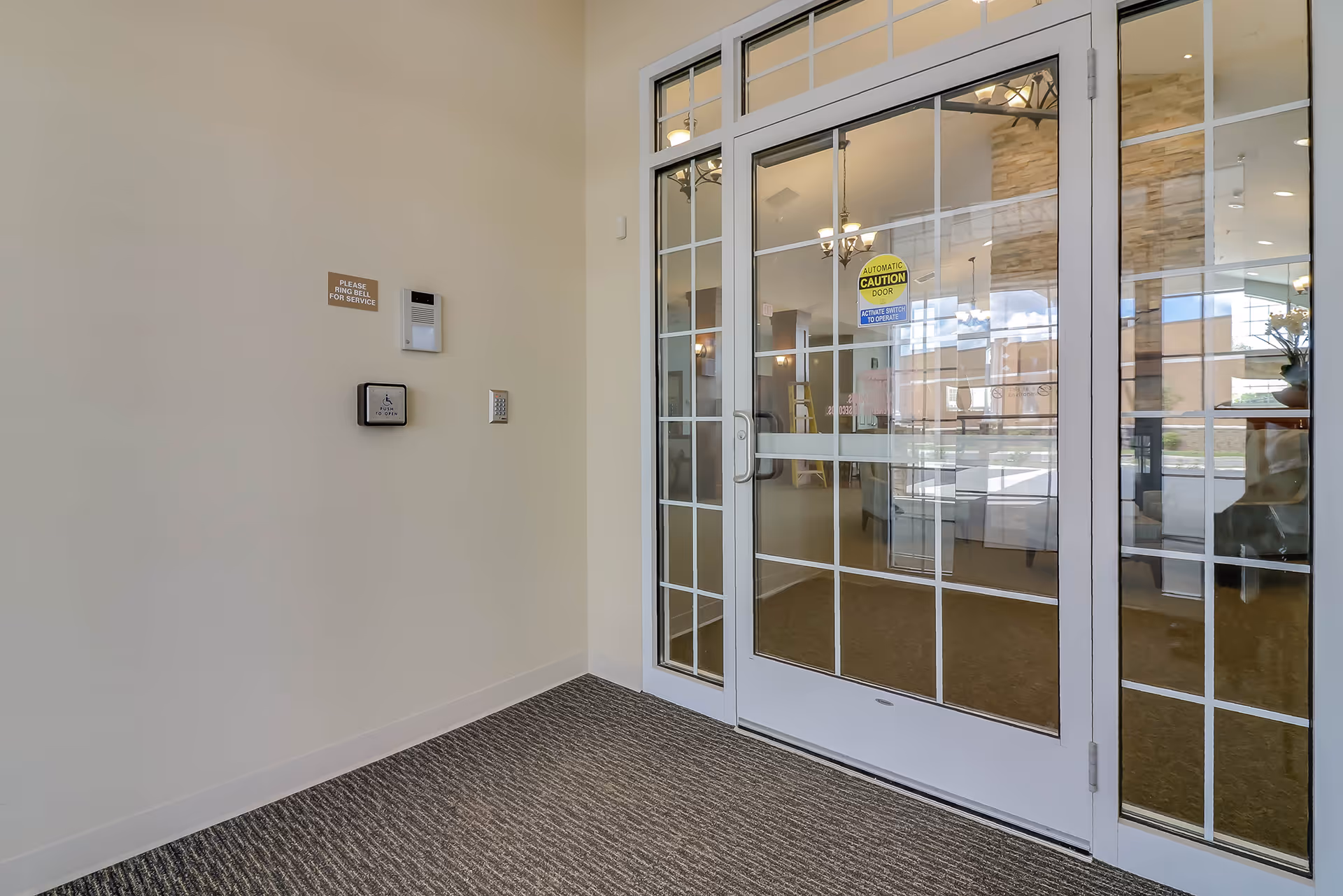 Glass double doors with window grids opening into a bright lobby, carpeted entry and wall-mounted intercom and service sign.