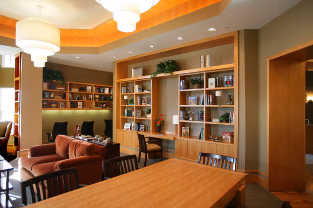 A cozy interior common area with wooden bookshelves filled with books and decorative items, a wooden table with chairs in the foreground, and comfortable seating including a brown sofa and black chairs. The room is warmly lit with ceiling lights and has a modern, inviting atmosphere.