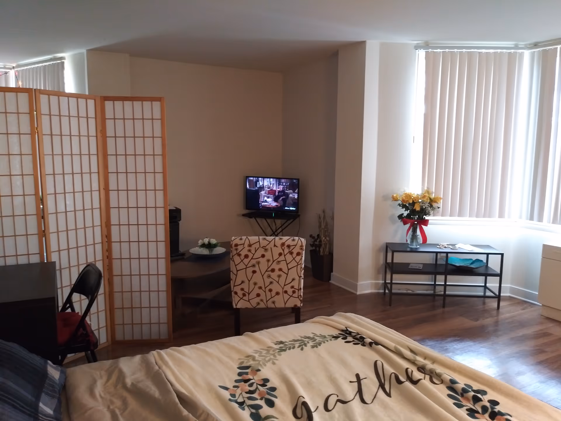 A cozy living space with a bed in the foreground covered with a blanket that says 'gather'. There is a patterned chair facing a small TV on a stand in the corner. A folding room divider and a black folding chair are on the left side. On the right, there is a small table with a vase of yellow flowers and vertical blinds covering the windows.