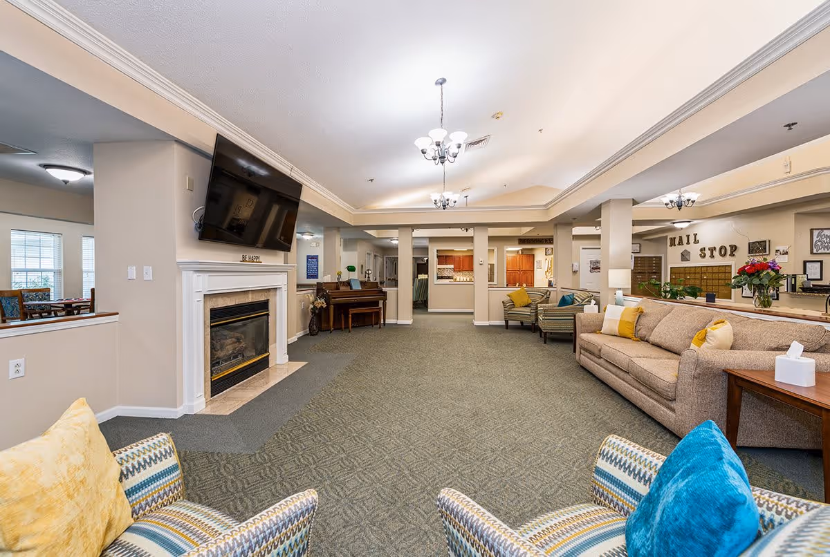 A spacious and well-lit living room area in a senior living facility with comfortable seating including a beige sofa with yellow and white pillows, patterned armchairs with colorful cushions, a fireplace with a mounted flat-screen TV above it, and a piano in the background. The room has carpeted flooring, beige walls, and ceiling lights with chandeliers. There is a mail stop area visible in the back right corner.