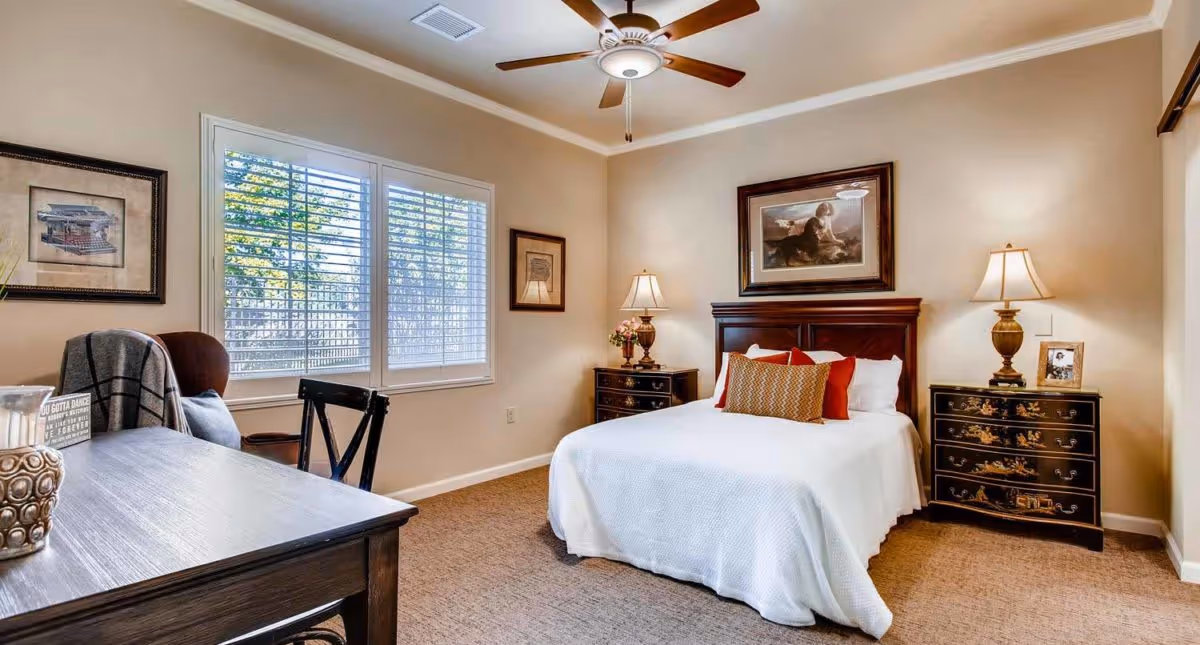 A cozy bedroom in a memory care community featuring a neatly made bed with white bedding and decorative pillows, two dark wooden nightstands with lamps on each side, framed artwork on the walls, a ceiling fan with light, a window with white shutters, and a wooden desk with a chair and decorative items.