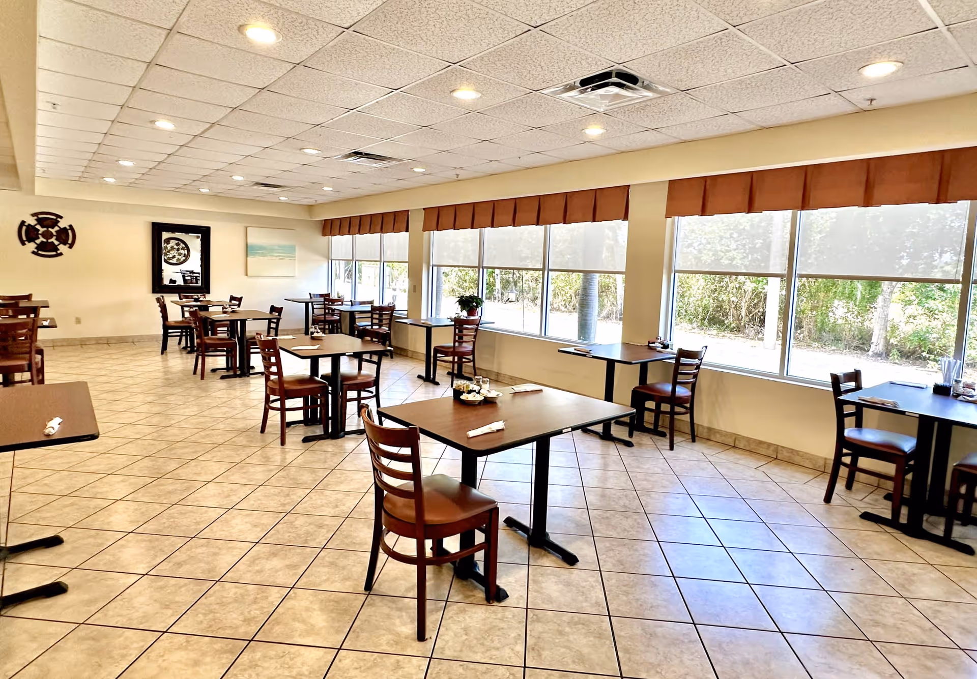 A bright dining room with multiple square wooden tables and chairs arranged neatly. Large windows along one wall allow natural light to fill the room, with a view of greenery outside. The floor is tiled, and the ceiling has recessed lighting. The room has simple wall decorations including a framed mirror and artwork.