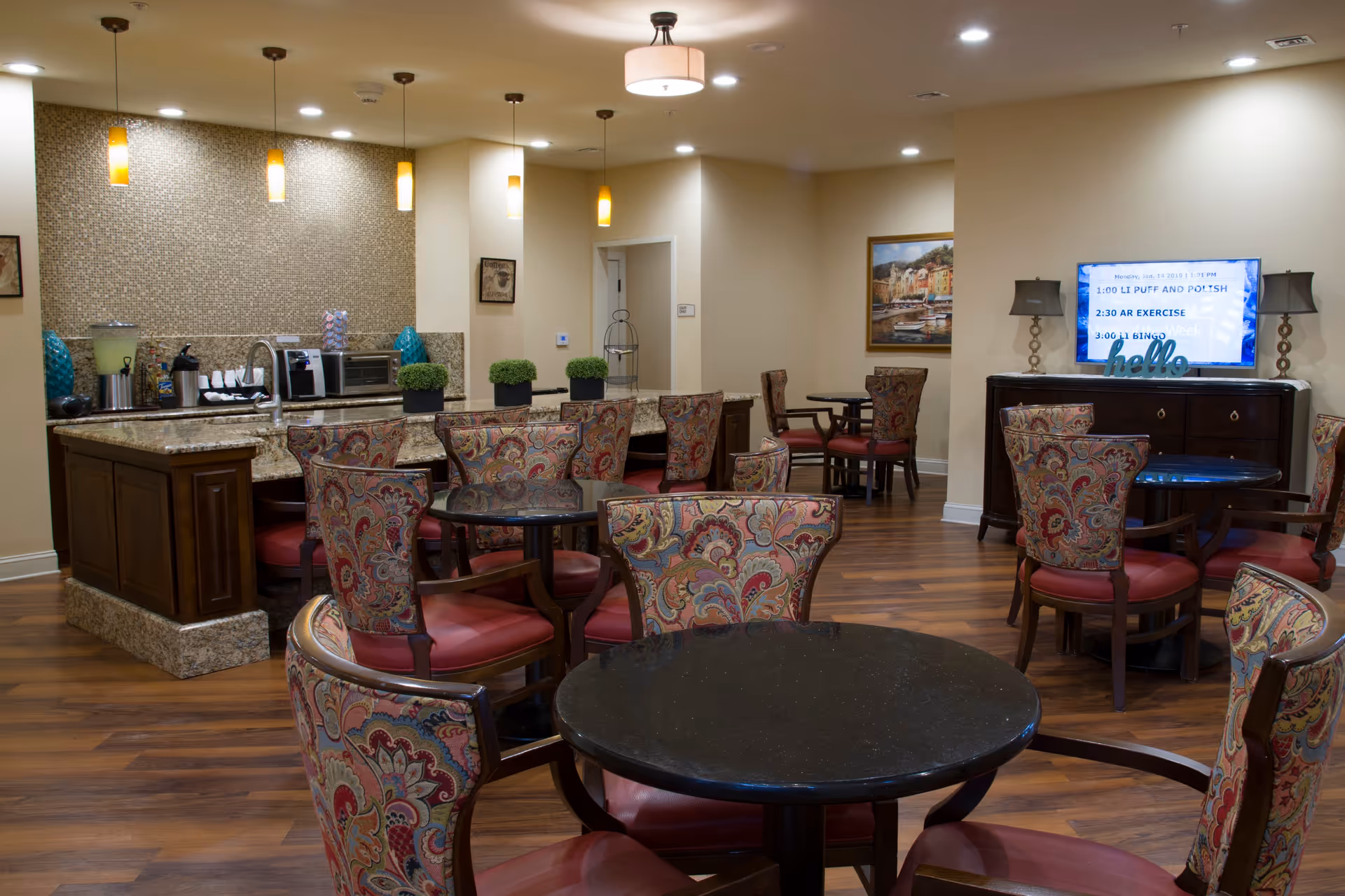 Communal dining/lounge area with round tables and patterned chairs, a granite-topped serving counter, and a wall-mounted TV.
