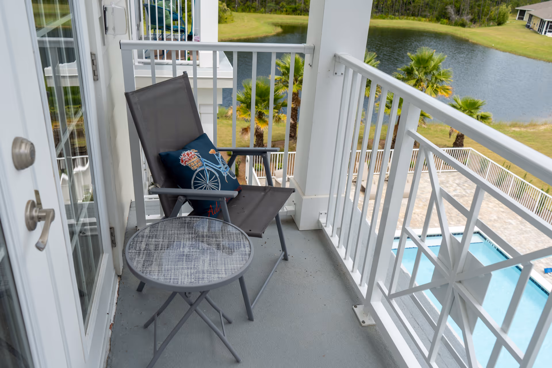 A small balcony with a gray folding chair featuring a decorative pillow with a bicycle and flowers design, and a round glass-top table. The balcony has white railings and overlooks a swimming pool, palm trees, and a pond with grassy areas.