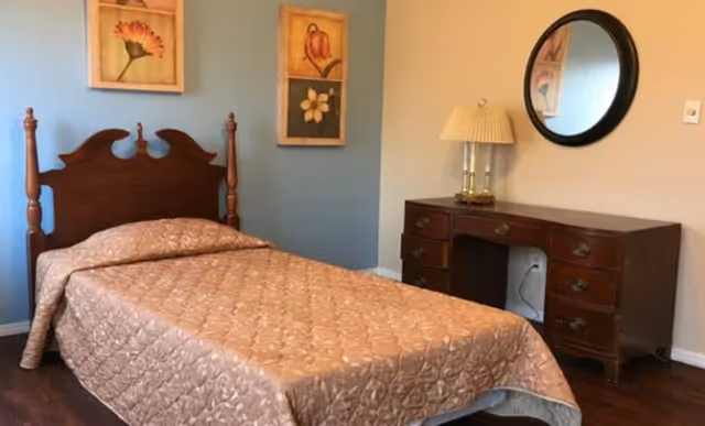 A simple bedroom with a single bed featuring a wooden headboard, a quilted bedspread, a dresser with a lamp and round mirror, and framed wall art.
