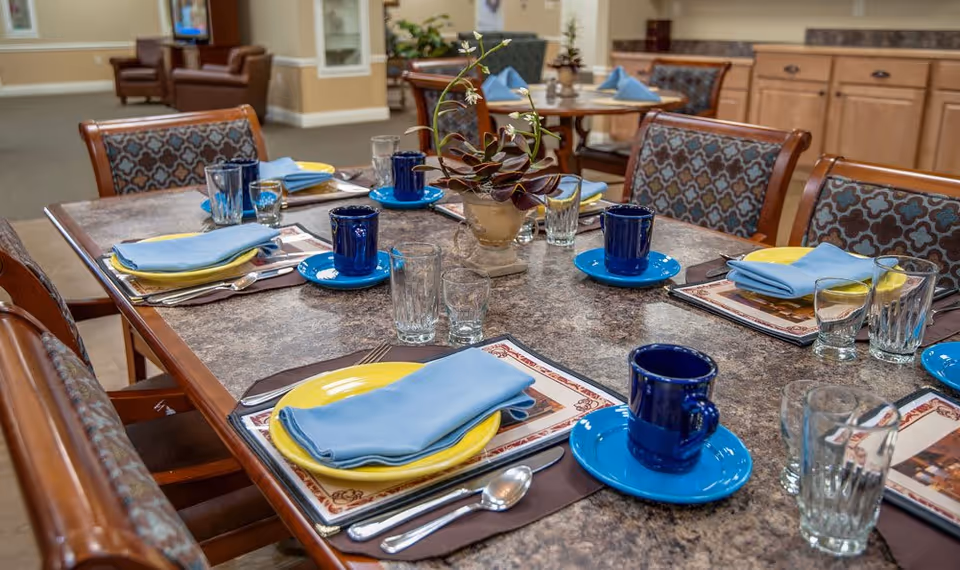 Communal dining table set with blue mugs, yellow plates, blue napkins, glassware, and a small plant centerpiece in an assisted living dining room.