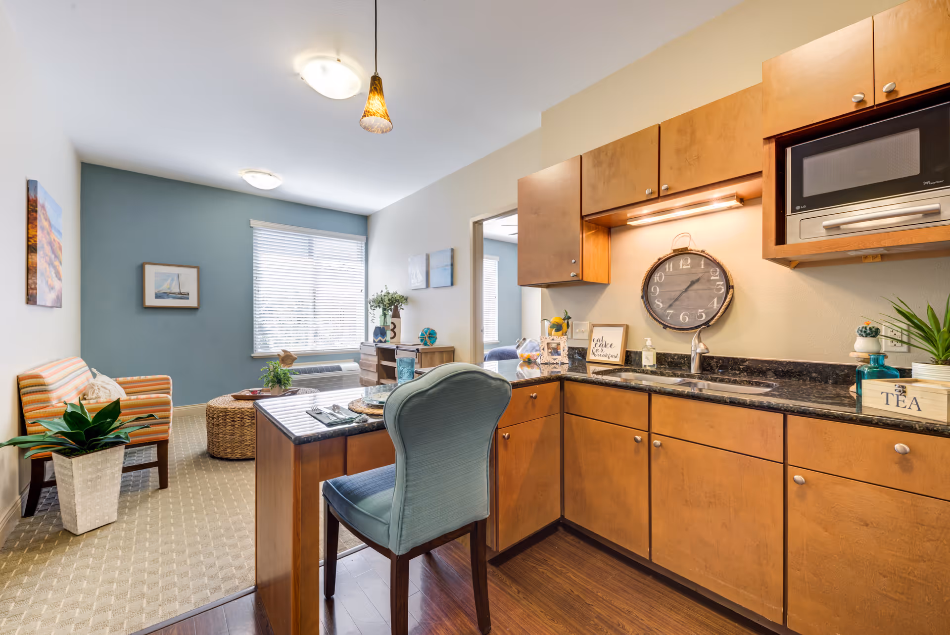 Open-plan kitchenette with wooden cabinets, granite countertop and bar seating opening to a small living area with chairs and a window.