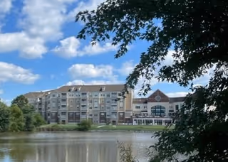 Multi-story senior living building viewed across a pond with trees framing the scene under a blue sky.