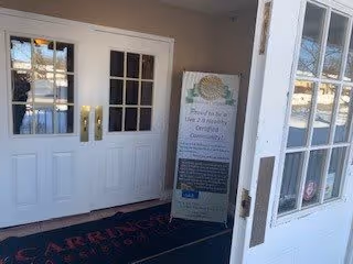 Entrance area with white double doors featuring glass panels. A standing signboard is placed near the doors, and a black mat with red text is on the floor. The area is well-lit with natural light coming through windows.