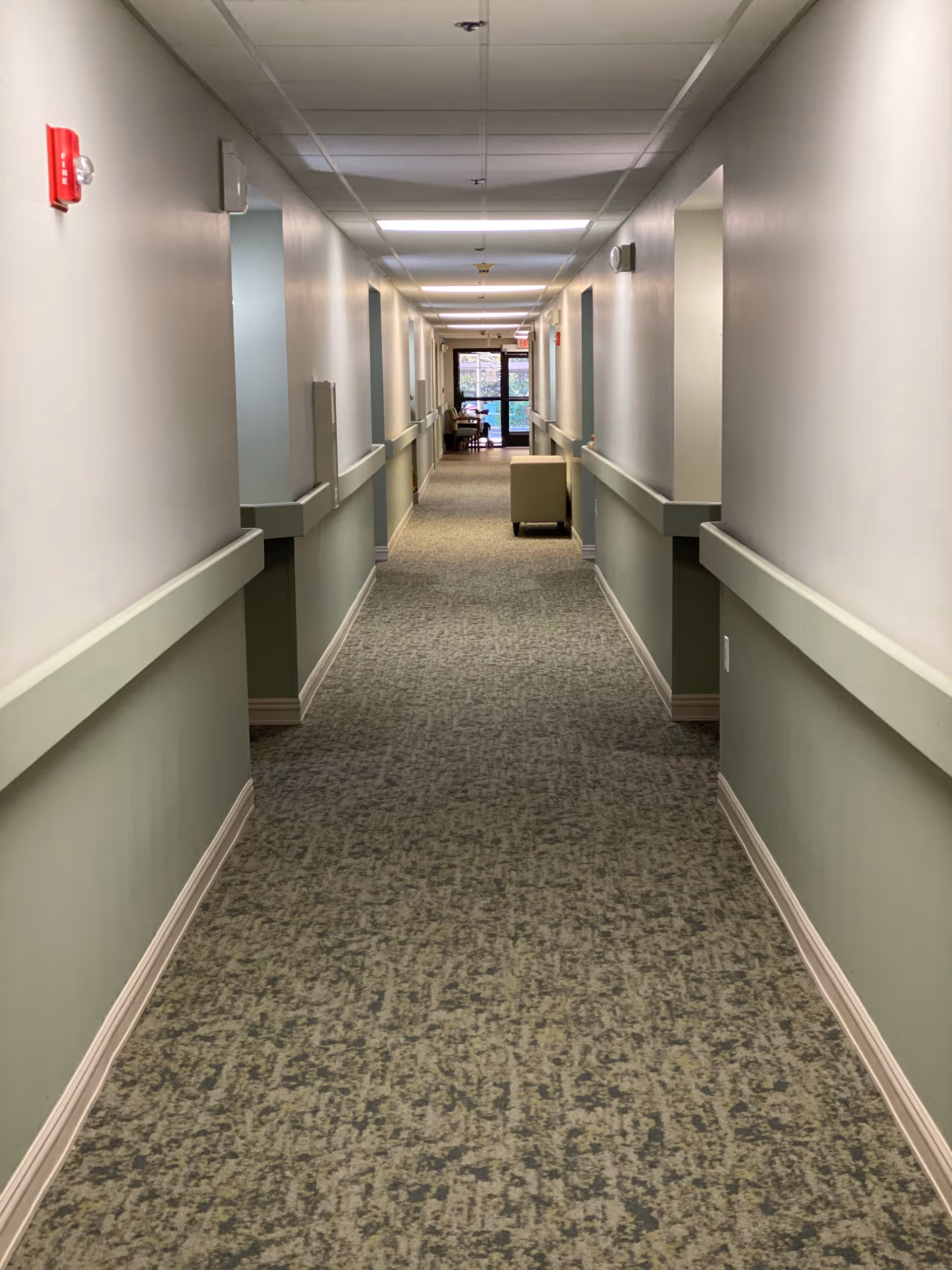 Long indoor hallway with light green lower walls and white upper walls, carpeted floor, handrails on both sides, ceiling lights, and a chair placed near the end of the corridor by a glass door.