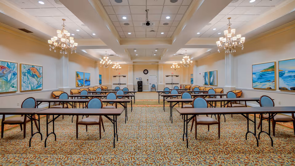 A spacious, well-lit meeting or dining room with multiple long tables and blue cushioned chairs arranged in rows. The room features chandeliers hanging from the ceiling, patterned carpet flooring, and colorful paintings of ocean scenes on the walls. There are double doors at the far end of the room and additional seating along the walls.