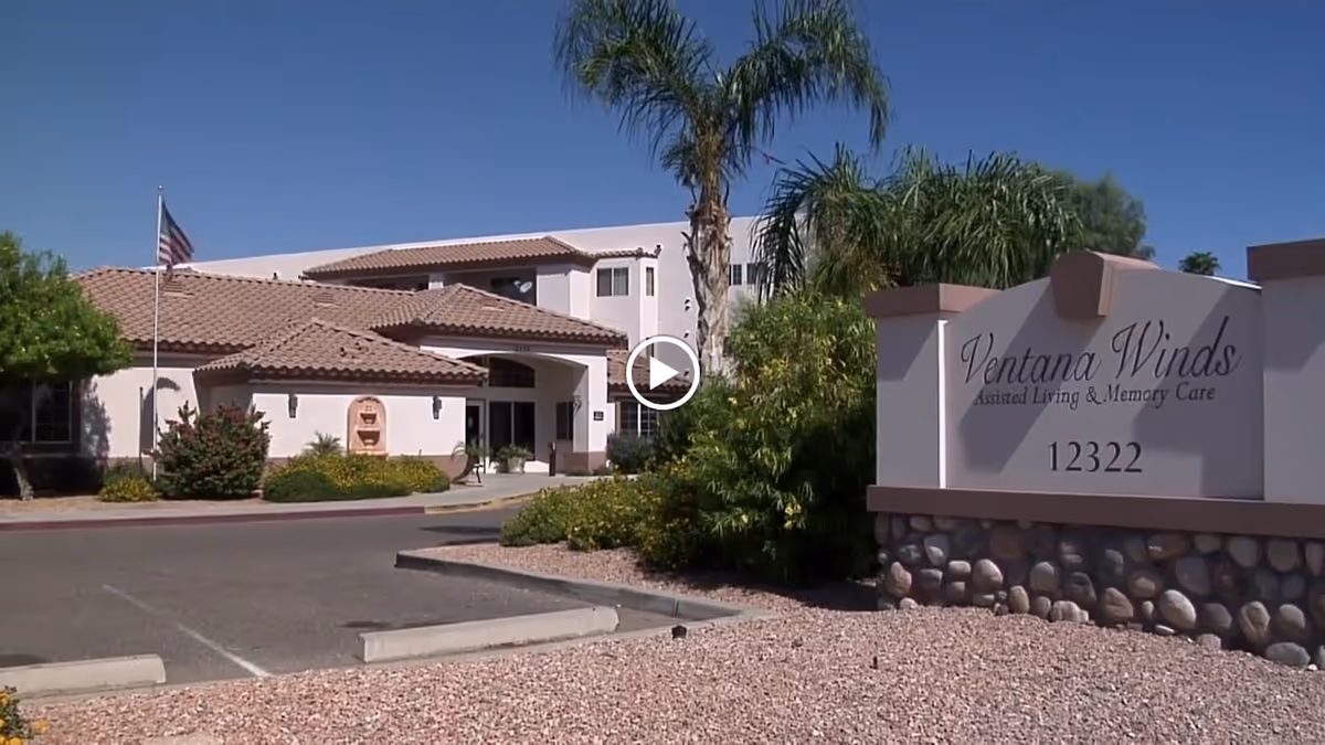 Exterior view of Ventana Winds Assisted Living & Memory Care facility with a clear blue sky, palm trees, and landscaped bushes. The building has a tiled roof and an American flag is visible near the entrance.