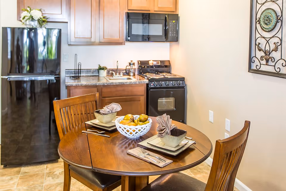 A small kitchen and dining area featuring a round wooden table set for two with square plates, bowls, napkins, and silverware. A white basket with fruit is centered on the table. The kitchen has wooden cabinets, a black refrigerator, a black microwave mounted above a black stove, and a small sink. A decorative wall hanging is visible on the right wall.