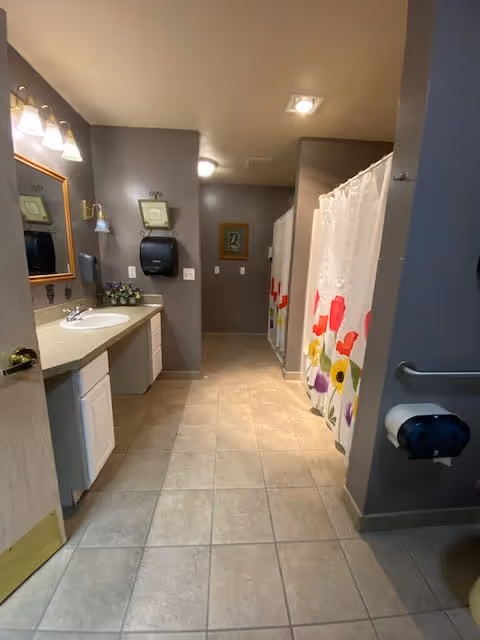 A clean and well-lit bathroom with beige tiled floor and gray walls. On the left side, there is a countertop with a sink, a mirror with lights above it, and a paper towel dispenser mounted on the wall. On the right side, there is a shower area with a colorful floral shower curtain. A toilet paper holder is visible on the right wall near the entrance.