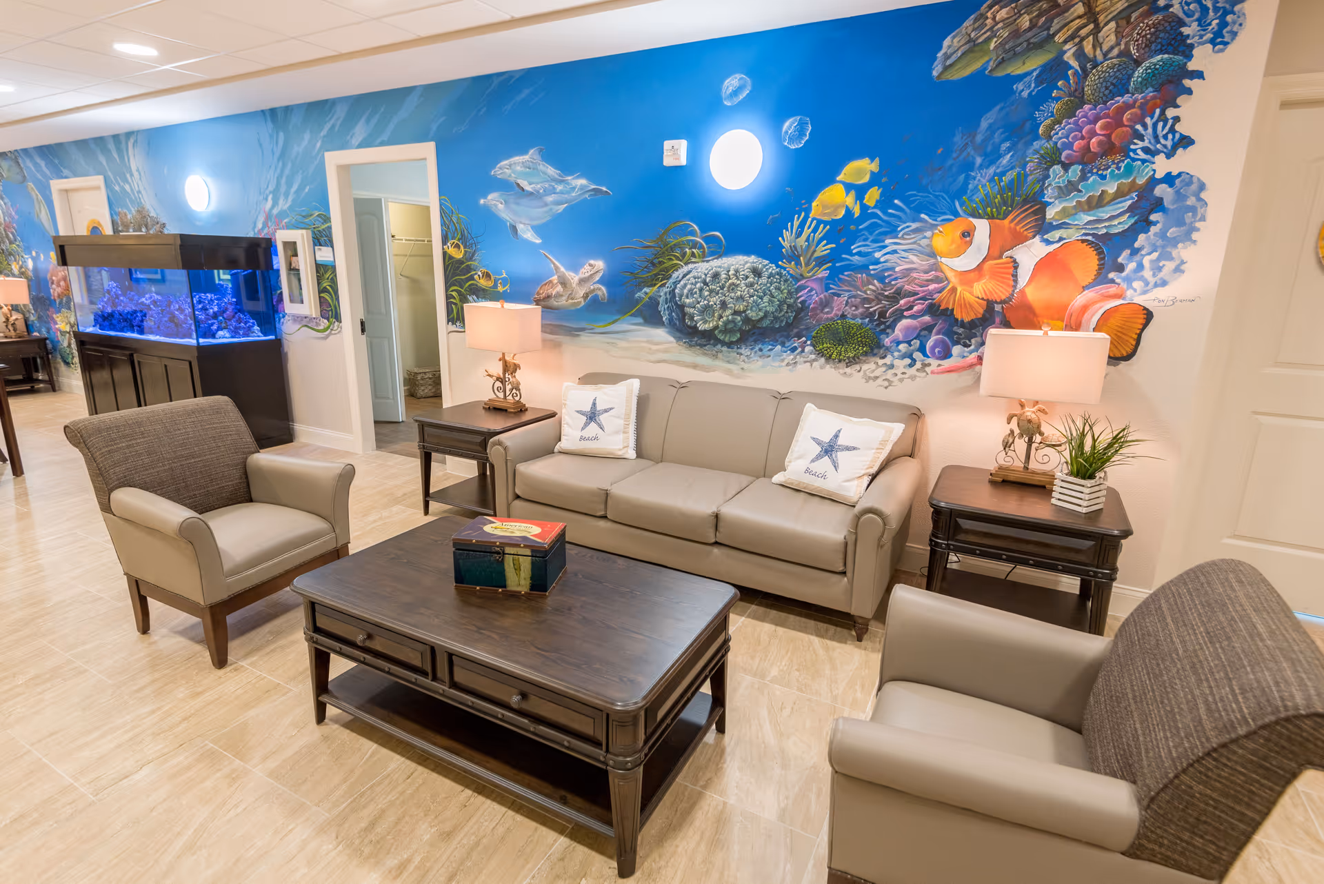 A cozy living room area with a beige sofa and two armchairs around a dark wooden coffee table. The walls feature a vibrant underwater mural with fish, coral, and sea turtles. There are two side tables with lamps and a large aquarium filled with colorful fish along one wall.