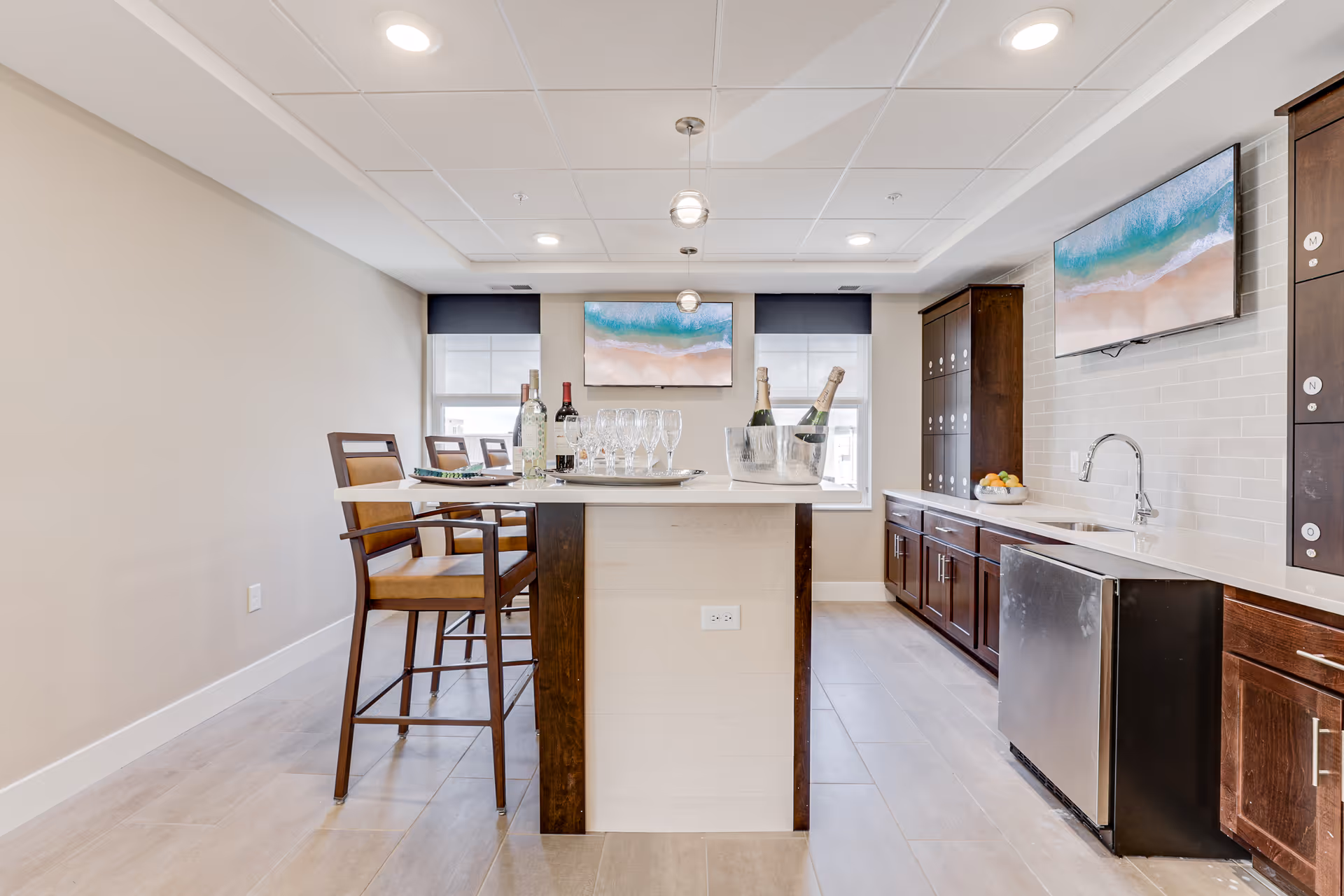 A modern kitchen area with a central island featuring two wooden bar stools. On the island, there are bottles of wine and champagne, glasses, and an ice bucket. The kitchen has dark wooden cabinets, a sink, a dishwasher, and two wall-mounted TVs displaying a beach scene. The room is well-lit with recessed ceiling lights and pendant lights above the island.