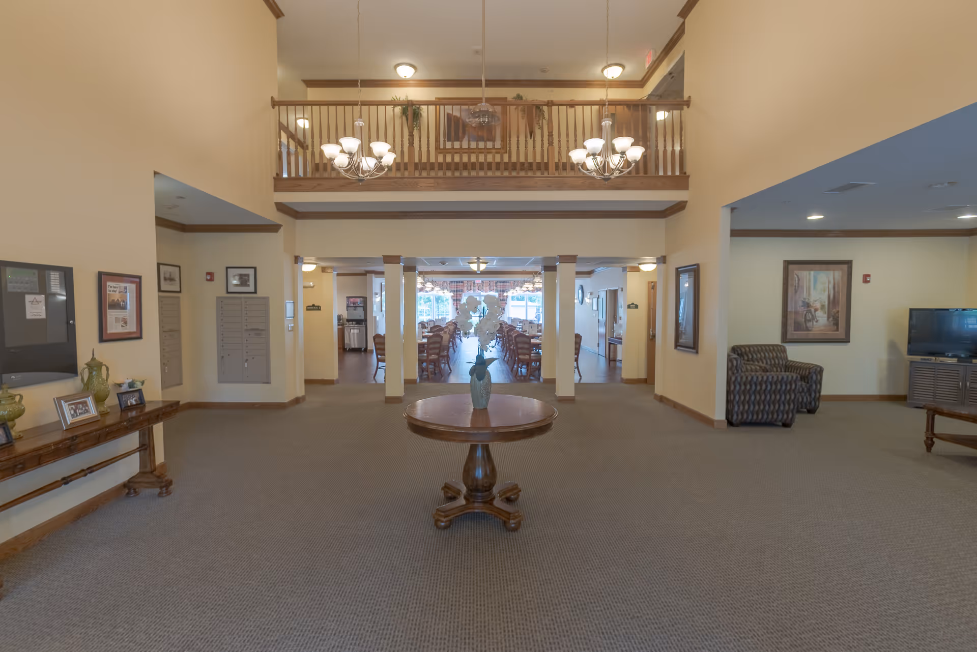 Spacious interior of a senior living facility with a central round wooden table holding a vase with white flowers. The room has beige walls, carpeted floor, and a high ceiling with two chandeliers. To the left, there is a wooden console table with decorative items and framed pictures. In the background, there is a dining area with multiple tables and chairs, and to the right, a seating area with an armchair and a TV on a wooden stand. A wooden railing balcony is visible above the dining area.