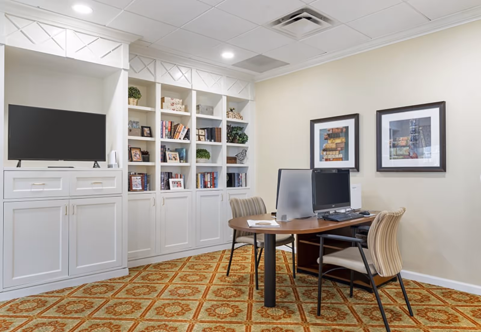 Well-lit common room with built-in white bookshelves and TV, a round table with computers and chairs, and framed artwork on the wall.