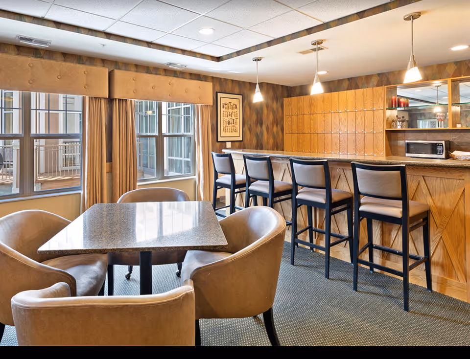 Common dining/lounge area with a table and upholstered chairs in the foreground and a bar counter with high stools along the right wall.