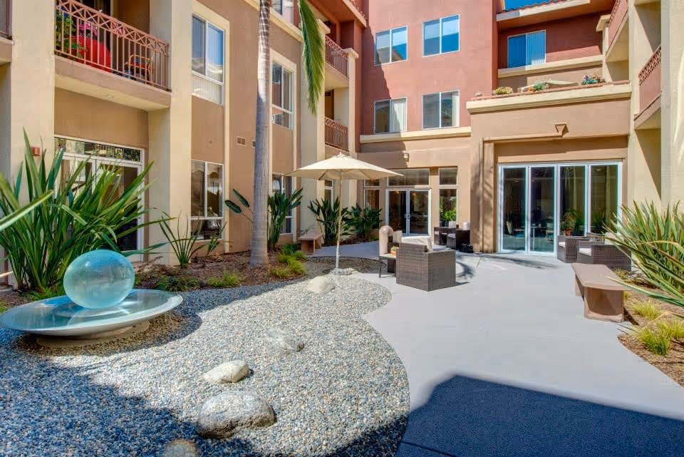 Outdoor courtyard area of a senior living facility with a decorative water feature consisting of a glass sphere on a metal dish surrounded by small rocks. The courtyard is bordered by a multi-story building with balconies and large windows. There are patio chairs, a table with an umbrella, benches, and various plants around the space.