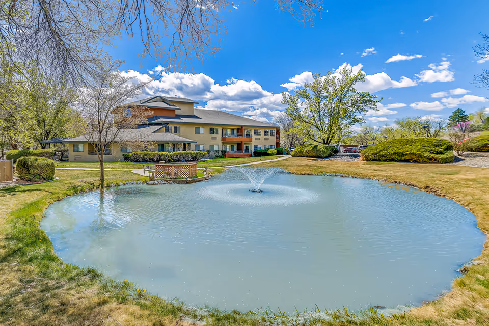 A senior living facility building with beige walls and a gray roof is visible behind a small pond with a water fountain in the center. The pond is surrounded by green grass and bushes, with trees around the area under a bright blue sky with scattered white clouds.