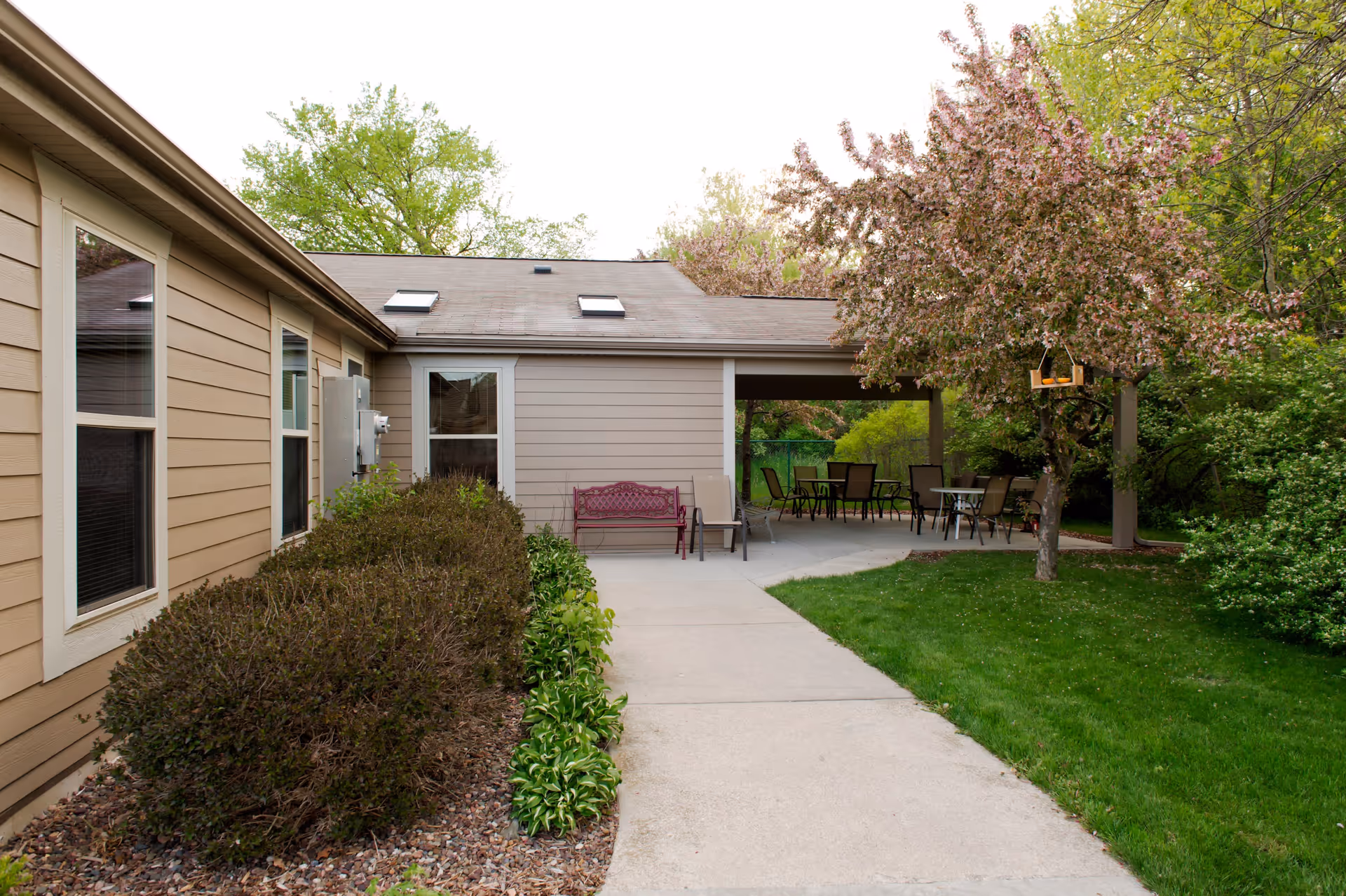 Outdoor patio area at Robins Landing at New Berlin with a concrete walkway leading to a covered seating area. There are several chairs and tables under the covered patio, surrounded by green grass, bushes, and a tree with pink blossoms.