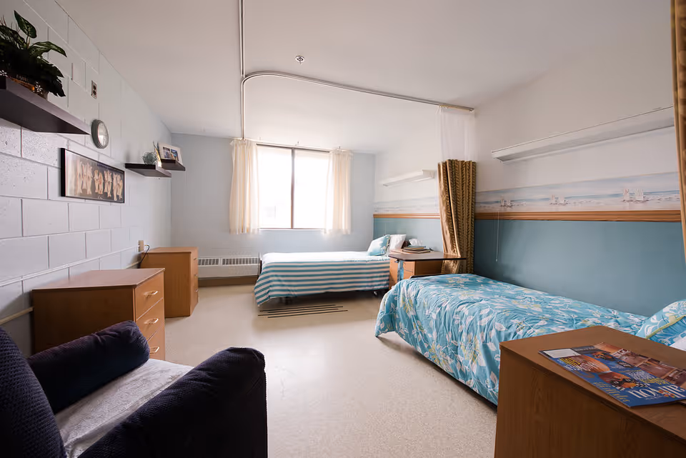 A bright, simple bedroom with two single beds, one with a blue and white striped bedspread and the other with a blue floral bedspread. The room has light blue walls with a beach-themed wallpaper border, a window with white curtains, wooden dressers, a small armchair, and shelves with decorative items.