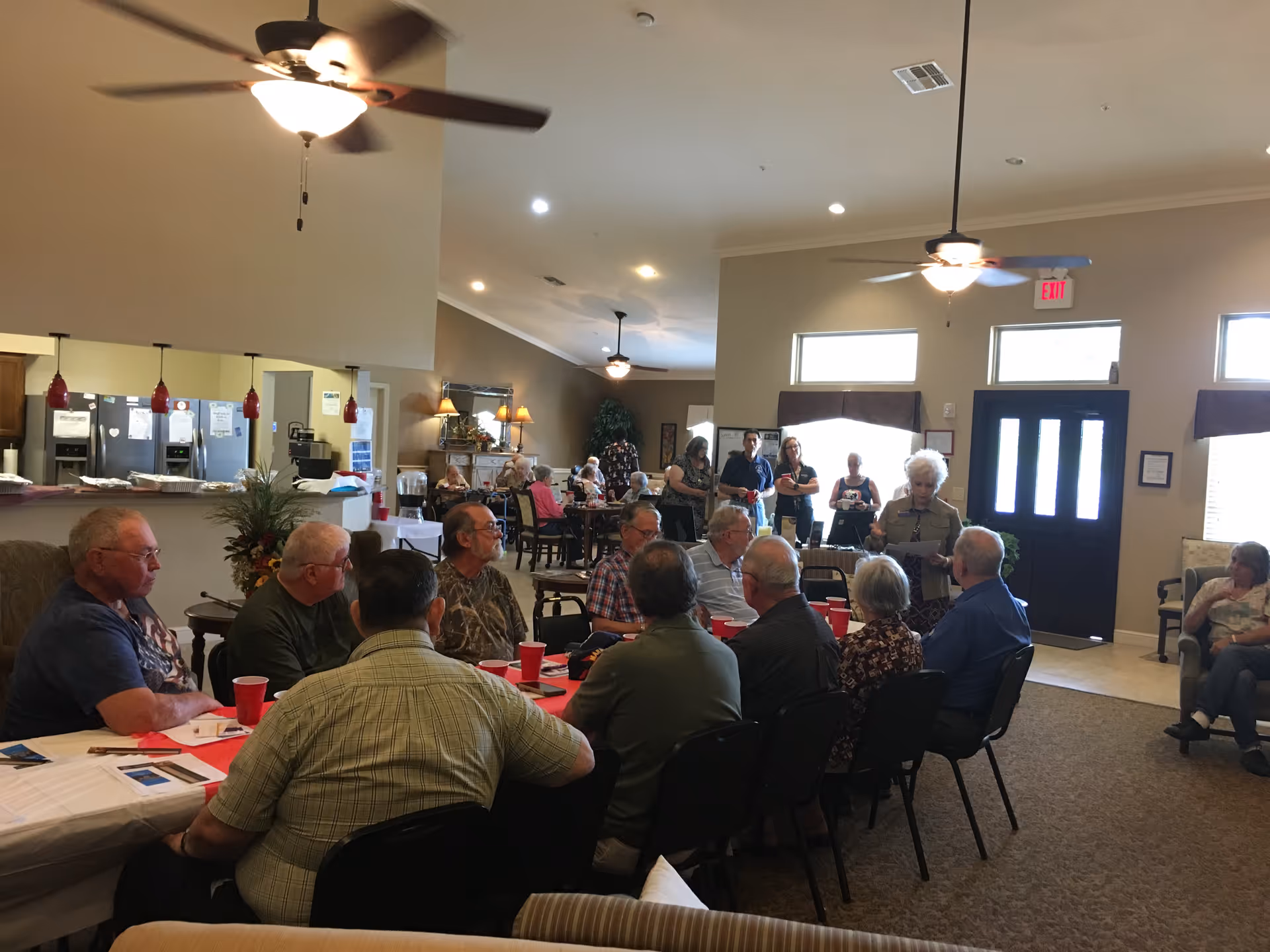 A group of elderly people seated around a table in a common area of an assisted living facility. Some people are standing and talking near the entrance door. The room has ceiling fans, large windows, and a kitchen area visible in the background.