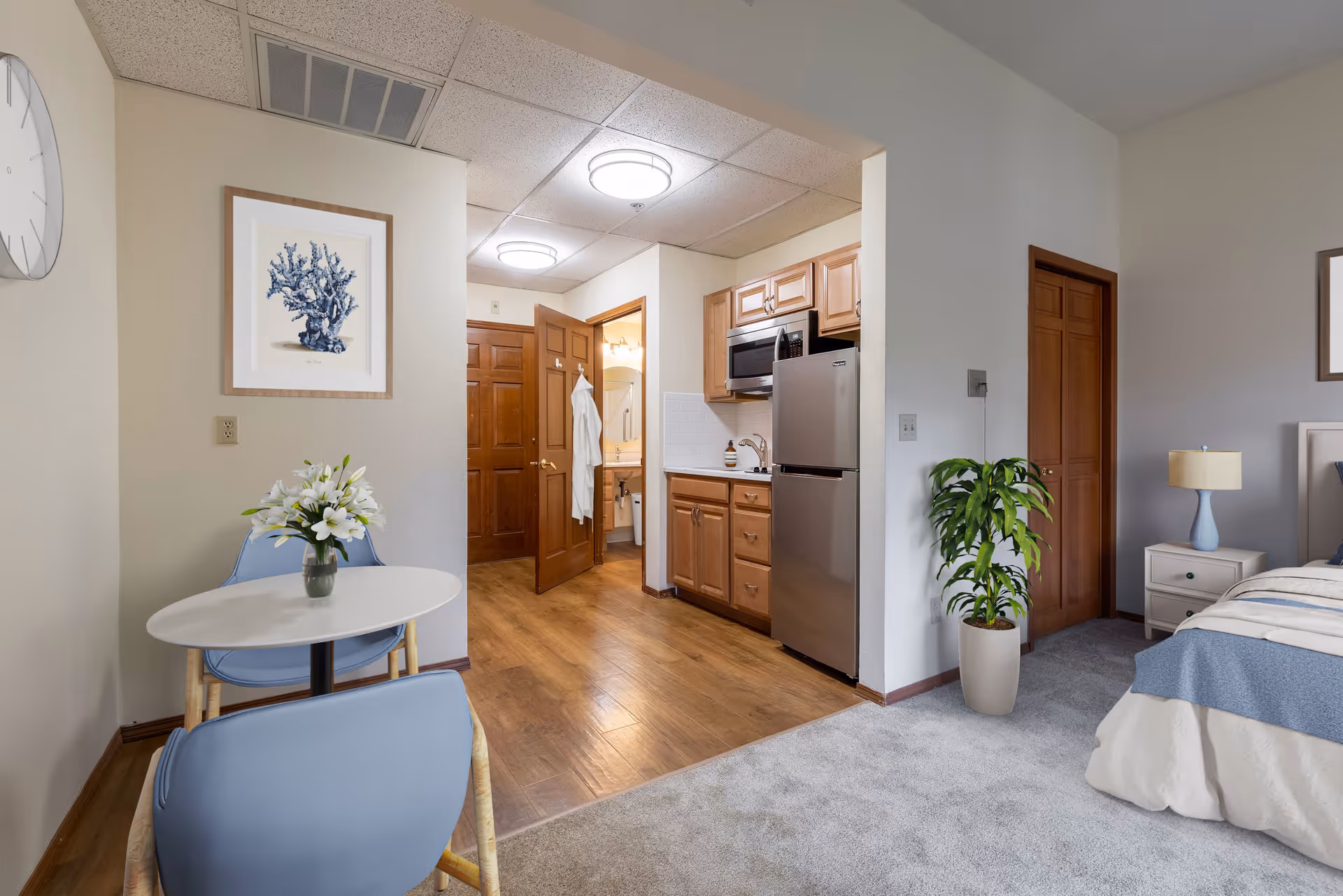 Interior view of a senior living residence showing a small dining area with a round white table and two blue chairs, a kitchenette with wooden cabinets, a stainless steel refrigerator and microwave, a doorway leading to a bathroom with a white robe hanging on the door, and part of a bedroom with a bed, nightstand, lamp, and a potted plant.