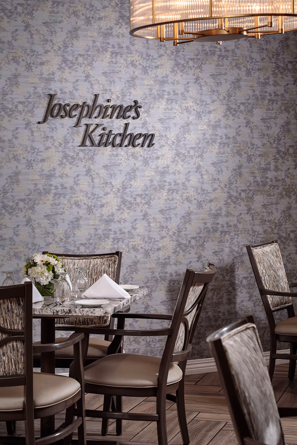 A dining area with a table set for four, featuring a floral centerpiece, folded white napkins, and glassware. The wall behind the table has a textured pattern with the words 'Josephine's Kitchen' mounted on it. Part of a modern chandelier is visible above.