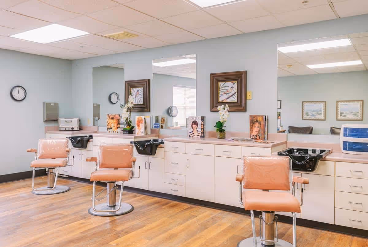 Interior of a senior living facility salon area with three peach-colored salon chairs in front of a long counter with sinks, mirrors, and cabinets. The room has light blue walls, wooden flooring, framed artwork, and a clock on the wall.