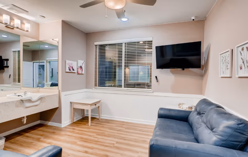 A cozy living room area with a blue leather couch, a small wooden side table, and a wall-mounted flat-screen TV. The room has light wood flooring, beige walls with white wainscoting, a ceiling fan with a light, and a large window with white blinds. There is a sink with a large mirror on the left side of the room, and framed floral artwork on the walls.