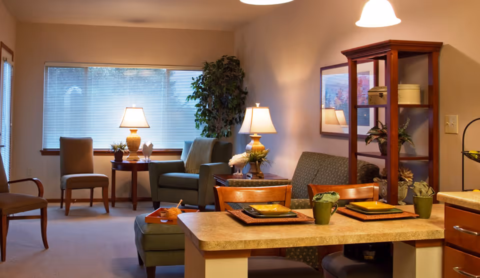 A cozy living room area with a large window covered by blinds, two table lamps on side tables, a green armchair, a patterned loveseat, and a wooden shelving unit with decorative items. In the foreground, there is a countertop with place settings including plates, cups, and napkins on woven placemats, along with wooden chairs.