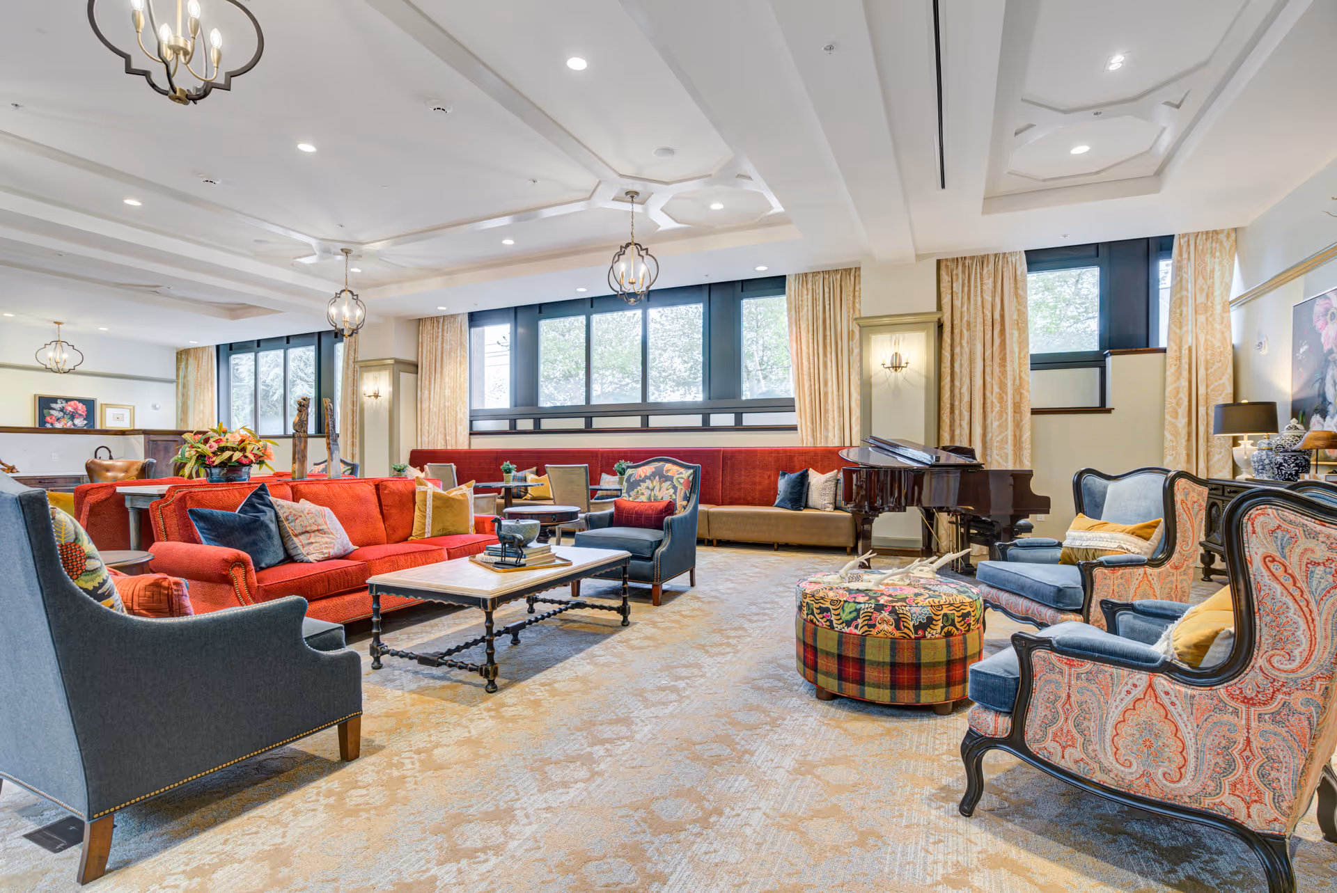 A spacious and elegantly furnished living room with a red sofa, multiple armchairs with patterned upholstery, a round plaid ottoman, a coffee table, and a grand piano near large windows with beige curtains. The ceiling features decorative molding and hanging light fixtures.