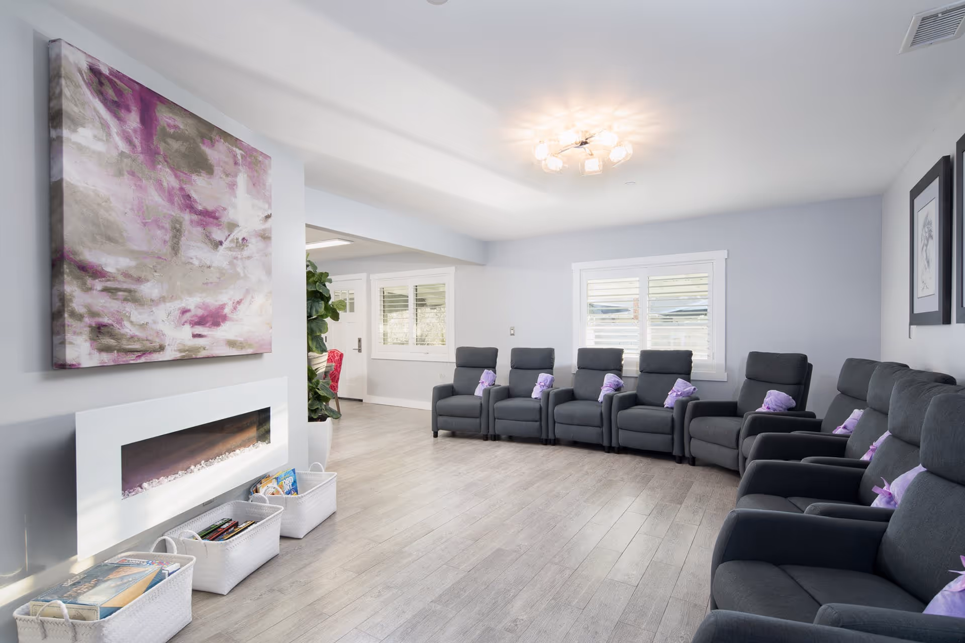 Bright communal lounge with a row of gray recliner chairs with purple pillows, a modern fireplace, wall art, and baskets of books.