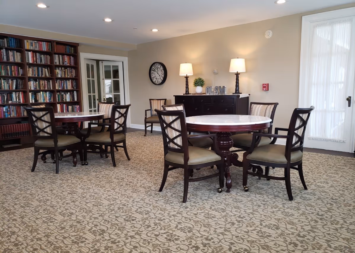 A cozy interior room with two round wooden tables, each surrounded by four cushioned chairs. There is a large bookshelf filled with books on the left side, a wall clock, and a dark wooden sideboard with two lamps and decorative items on top. The room has patterned carpet flooring and light-colored walls with a door covered by sheer curtains on the right.