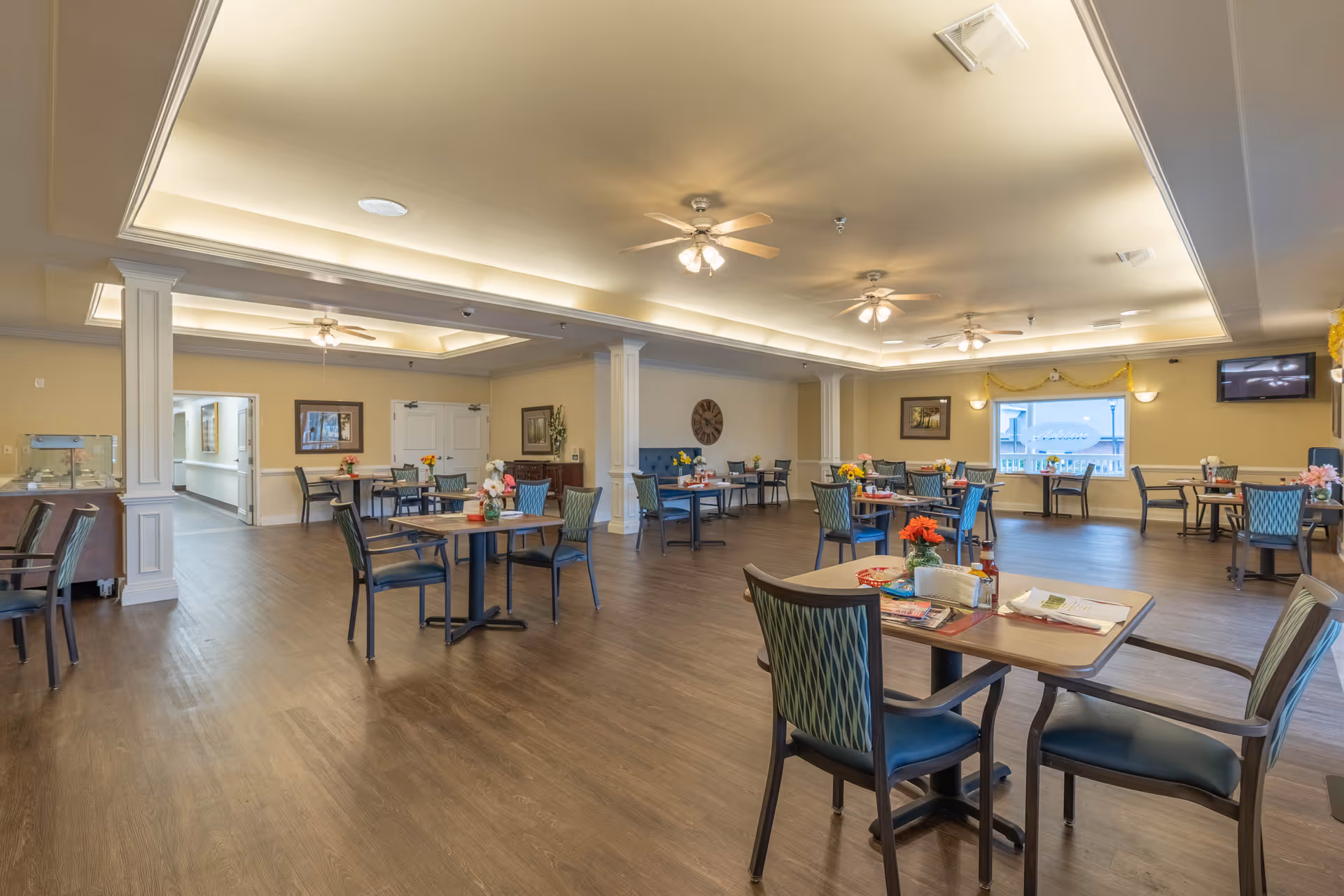 A spacious dining room with multiple tables and chairs arranged neatly. The room features a wooden floor, ceiling fans with lights, decorative columns, and soft yellow walls. Each table has small flower arrangements and condiments. There are windows letting in natural light and framed artwork on the walls.