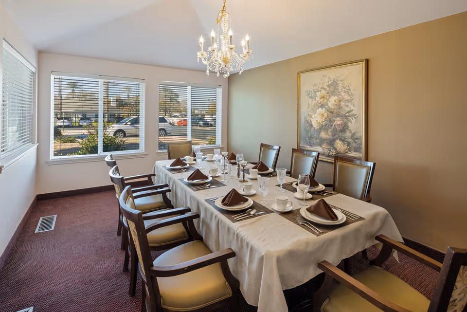 A dining room with a long table set for eight people. The table is covered with a white tablecloth and has brown napkins, white plates, cups, and glassware. There are eight wooden chairs with cushioned seats around the table. A chandelier hangs from the ceiling above the table, and a large floral painting is on the beige wall. Three large windows with blinds allow natural light to enter the room, showing a parking lot and palm trees outside.