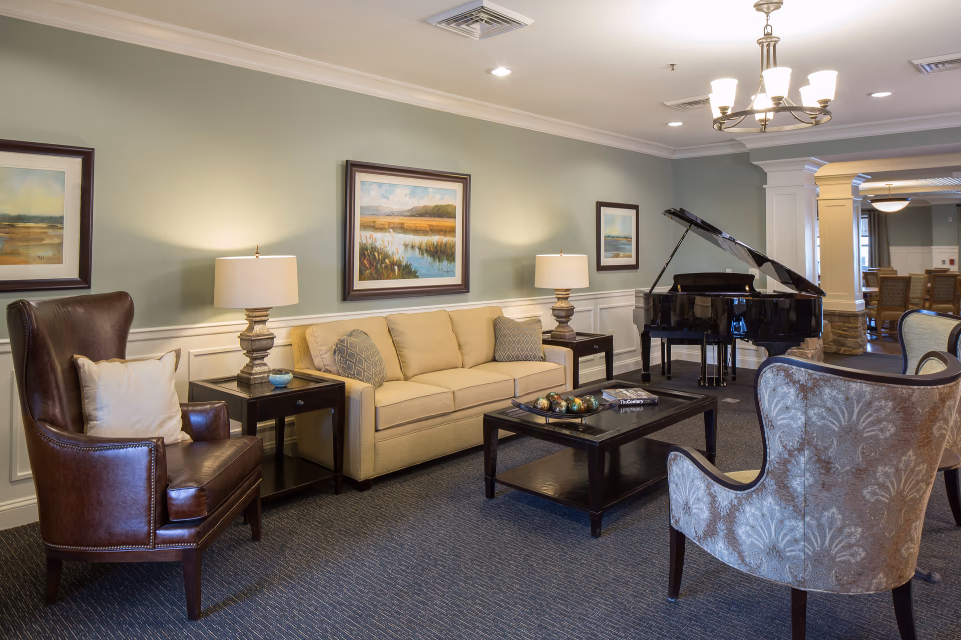 A cozy senior living facility lounge area with a beige sofa, two patterned armchairs, a dark leather armchair, two side tables with lamps, a black coffee table with decorative items, and a grand piano in the background. The walls are painted light green with white wainscoting and decorated with framed landscape paintings.