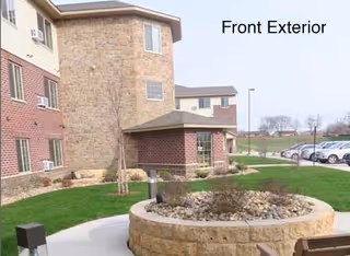 Exterior view of a senior living facility showing a brick and stone building with multiple windows, a landscaped area with grass, a circular stone planter, a small covered entrance, and a parking lot with cars in the background under a clear sky.