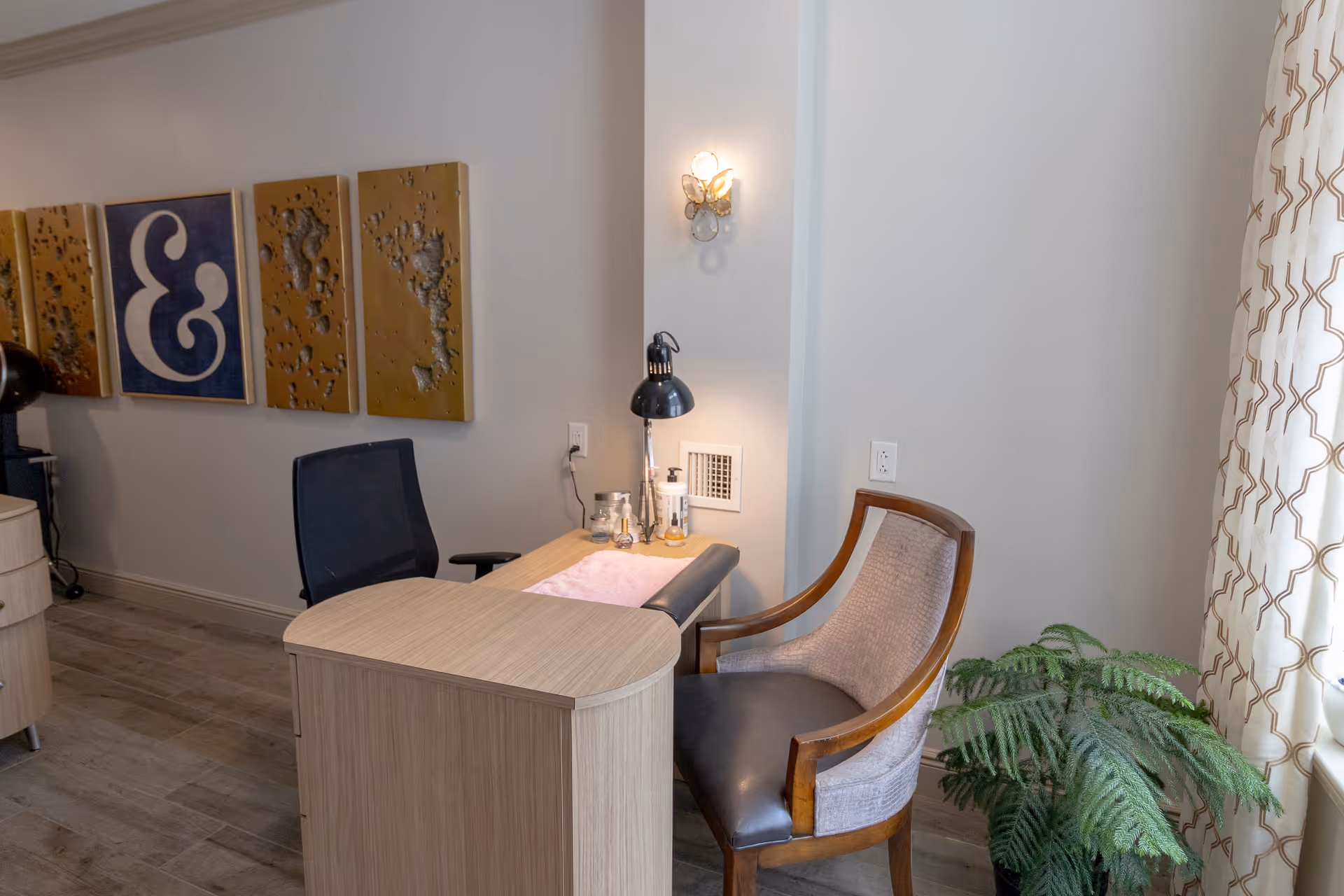 A small office or consultation area with a light wood desk, a black office chair, and a cushioned wooden armchair. The desk has a black desk lamp, a pink towel, and some small bottles. On the wall behind the desk are four decorative panels, one featuring a large ampersand symbol. A potted green plant is near a window with patterned curtains.