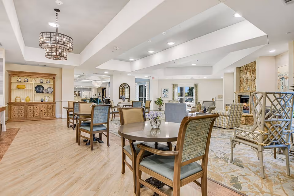 A spacious and well-lit common area in a senior living facility featuring multiple round tables with wooden chairs, a cozy seating area with armchairs near a stone fireplace, decorative shelving with plates and vases, and large windows allowing natural light to fill the room.