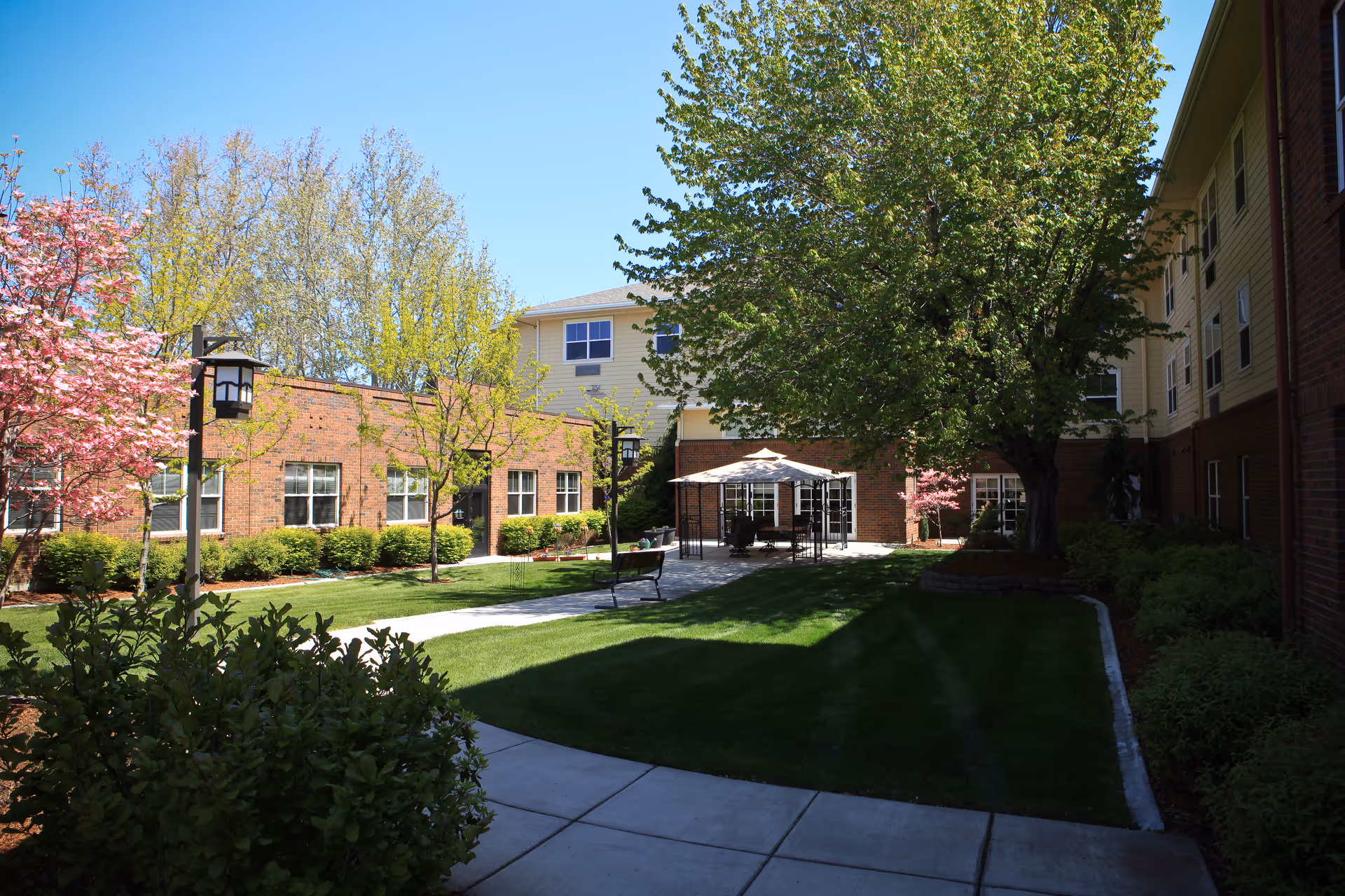 Outdoor courtyard area at Hillcrest At Summitview with green grass, trees including one with pink blossoms, a paved walkway, benches, and a gazebo with seating underneath. The courtyard is surrounded by a multi-story brick and siding building under a clear blue sky.