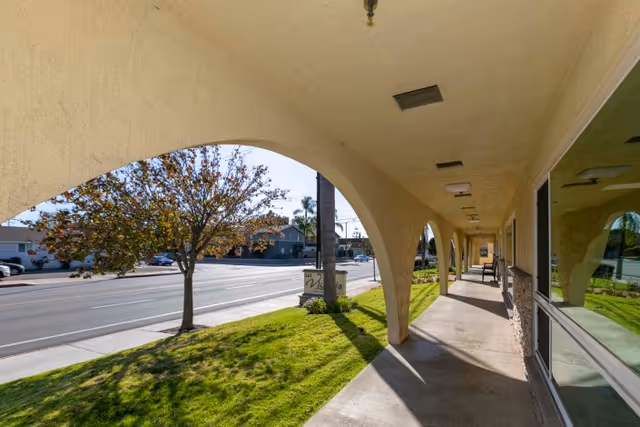 Covered arched walkway along the front of a building overlooking a street with grass and trees.