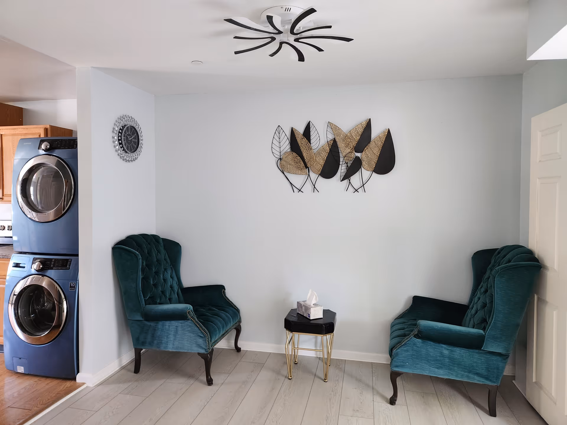 A small seating area with two teal upholstered armchairs facing each other and a small black and gold side table with a tissue box on it. On the wall above the table is a decorative metal wall art featuring black and gold leaves. To the left, there is a stacked washer and dryer unit partially visible, and a wall clock is mounted on the adjacent wall. The floor is light-colored wood, and the walls are painted light gray. A modern black ceiling fan is mounted on the ceiling.