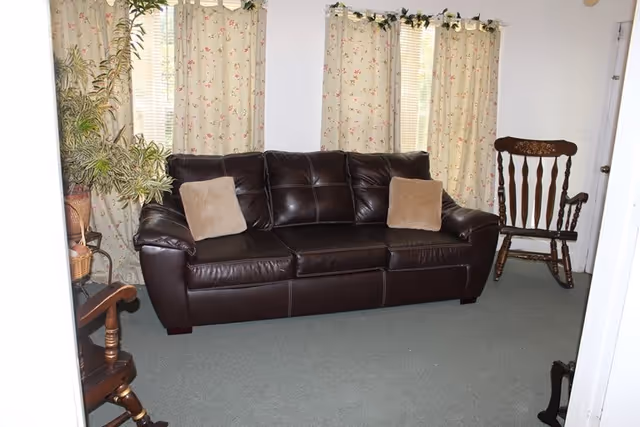 A cozy living room with a dark brown leather sofa adorned with two beige cushions, floral-patterned curtains covering the windows behind the sofa, a wooden rocking chair to the right, and a small wooden side table with a potted plant on the left.