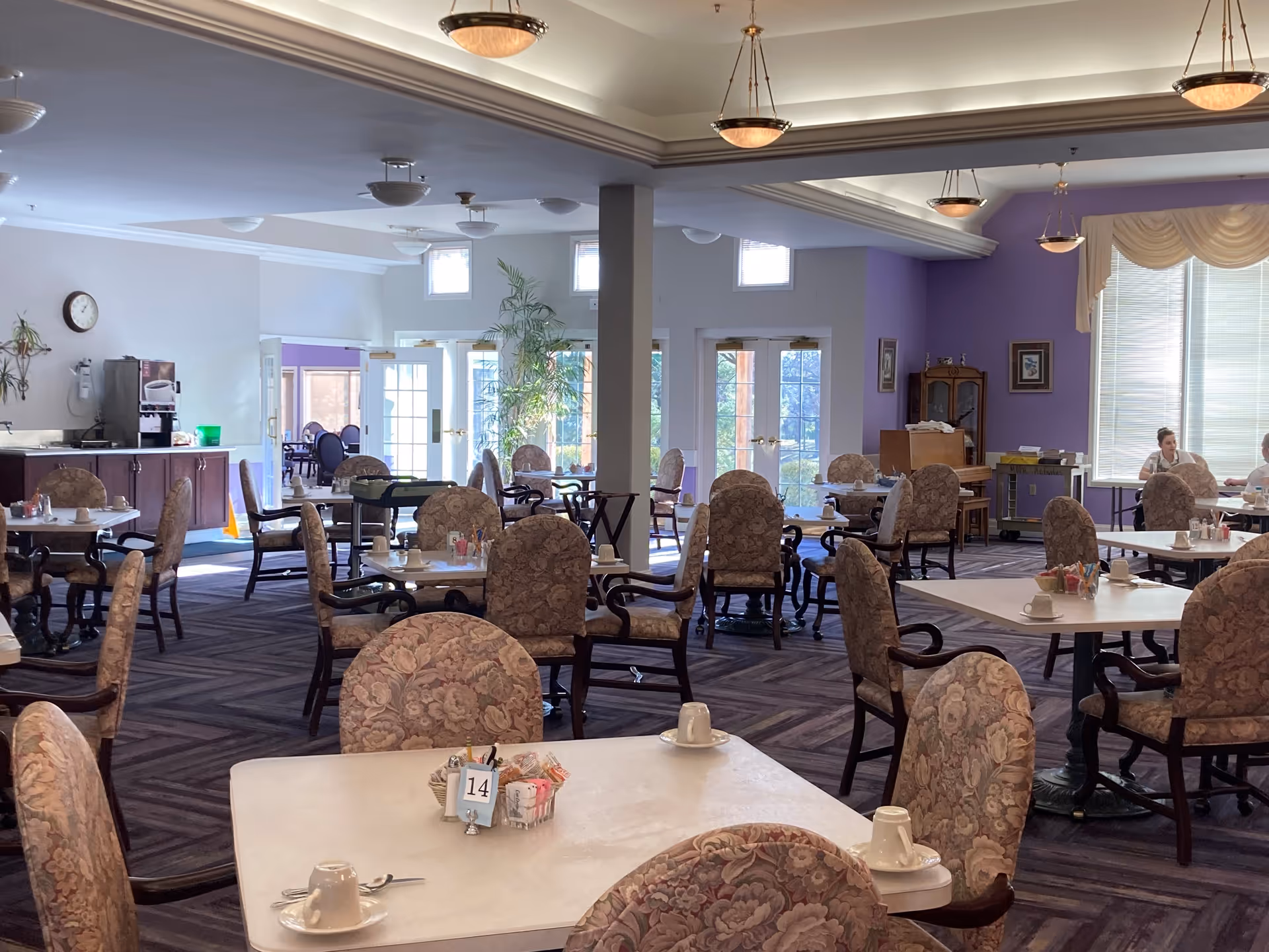 A spacious dining room with multiple tables and floral upholstered chairs arranged neatly. Each table has white cups and saucers, utensils, and condiment holders. The room has large windows with blinds, purple accent walls, and ceiling lights. Two people are seated at a table near the window on the right side of the room.