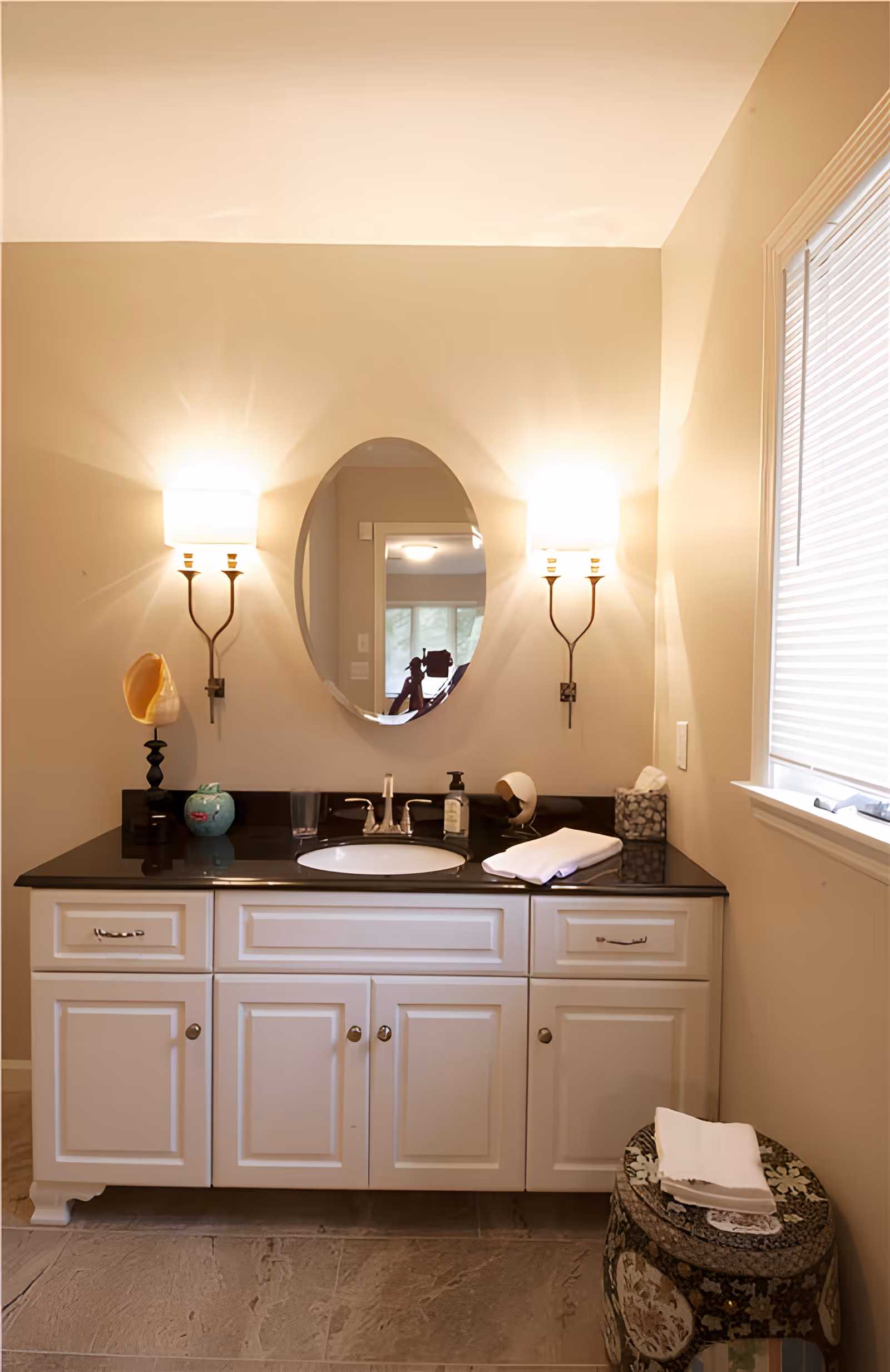 A bathroom vanity with a black countertop and white cabinets. An oval mirror is mounted on the wall above the sink, flanked by two wall sconces with white shades. On the countertop, there are decorative items including a shell, a small blue vase, a soap dispenser, a glass, and folded white towels. To the right, there is a window with blinds and a small decorative stool with folded towels on it.