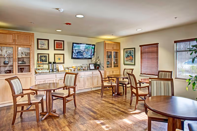 A bright dining area with wooden floors and several round wooden tables, each surrounded by cushioned chairs with striped upholstery. The room features light-colored wooden cabinets and a countertop with coffee and snack items. A flat-screen TV is mounted on the wall above the counter, and framed artwork decorates the walls. Two windows with wooden blinds allow natural light to enter the space.
