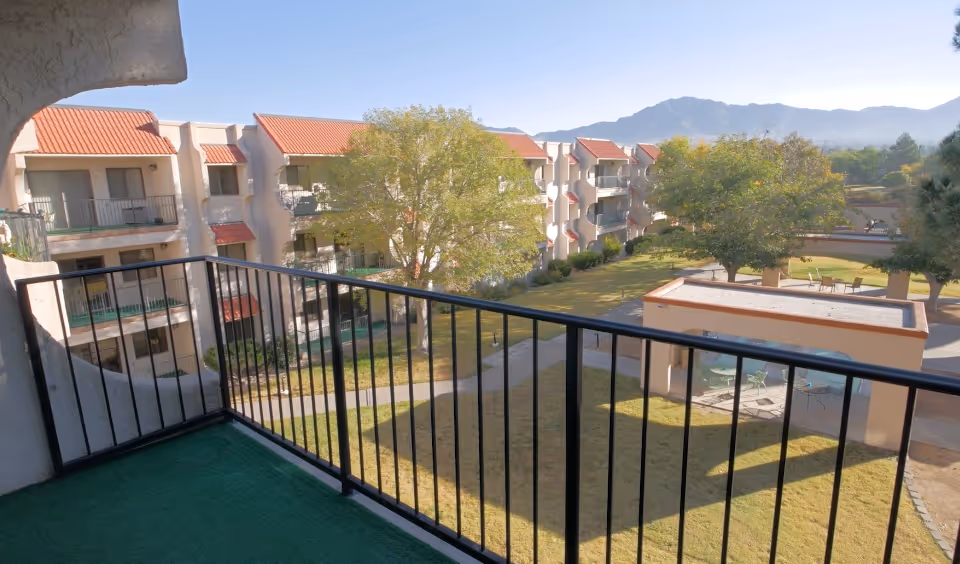 View from a balcony with black metal railing overlooking a courtyard with grass, trees, and outdoor seating areas. Surrounding the courtyard are multi-story buildings with balconies and red-tiled roofs. Mountains are visible in the background under a clear sky.
